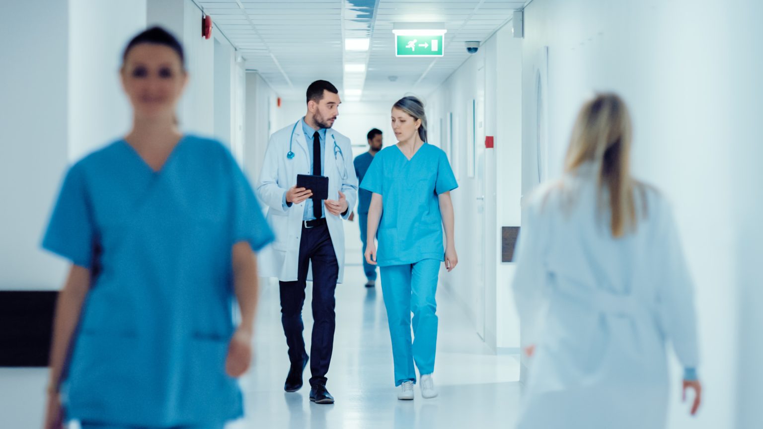 Surgeon and Female Doctor Walk Through Hospital Hallway, They Co